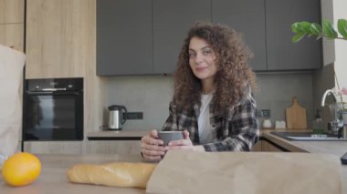 Gülümseyen kıvırcık kadın alışverişten sonra sıcak içeceğin tadını çıkarıyor. Huzurlu Sabah Anı Rahat Yaşam Tarzı, Ev Konforu, Ev İçi Sıcaklığı ve Bakkal Alışkanlığı Sonrası Kişisel Bakım