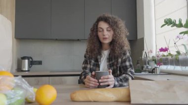 Kıvırcık kadın alışveriş yaptıktan sonra mutfakta bir fincan çayla hayal kuruyor. Parlak Modern Mutfakta Huzurlu Rahatlama Anı, Sakin Yaşam Tarzı Yansıtan Ev Atmosferi