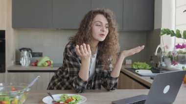 Sıcak mutfak çalışma odasında öğle yemeği molası sırasında görüntülü konuşan bir kadın. Parlak modern ev ortamında, meyvelerle çevrili taze salatanın tadını çıkarırken, uzaktan iletişimle meşgul olunuyor.