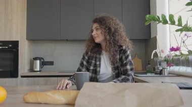 Mutfak masasında tablet açan genç bir kadın var. Rahat bir sabah sahnesinde esmer bir kadın sıcak bir evde çay içerken yemek tariflerini ya da haberleri kontrol etmeye hazırlanıyor.