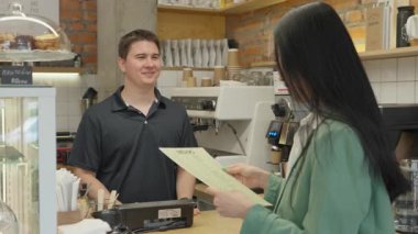 Müşteri, modern kafe tezgahında menü okurken kahve sipariş ediyor. Dostça bir etkileşim, her günün modaya uygun kahve mekanını sıcak atmosfer ve doğal detaylarla yakalar.