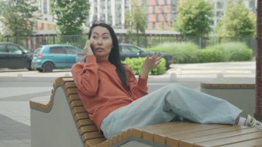 Modern Urban Bench 'te uzanırken telefonda konuşan rahat kadın. Şık şehir ortamında sıradan bir sohbet, kendine güvenen bir ifadeyle eğlenen genç bir bayan.