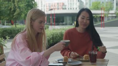 Güneşli bir günde açık kafede kahve içip sohbet eden iki kadın. İçkiler ve pastalarla masada oturan, gülümseyen ve sohbet eden modern şehir manzaralı rahat bayan arkadaşlar.