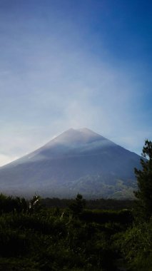 Capture the awe-inspiring view of Semeru Mountain from its foothills, where volcanic ash billows into the sky every minute. Surrounded by vibrant blue skies and diverse trees, clipart