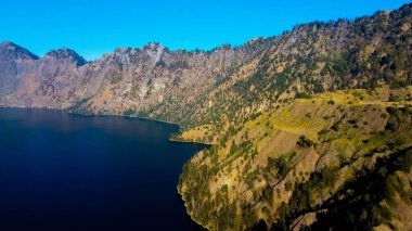Rinjani 'nin zirvesinden bir hava videosu aşağıdaki muhteşem Segara Anak Gölü' nü gösteriyor. Canlı mavi gökyüzü ve bulutlarla çevrili gölün dingin güzelliği dramatik manzaraya karşı duruyor..