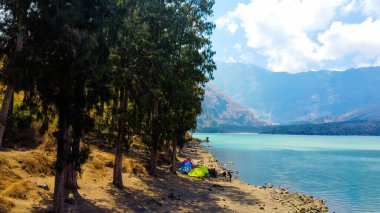 Rinjani 'nin zirvesinden bir hava videosu aşağıdaki muhteşem Segara Anak Gölü' nü gösteriyor. Canlı mavi gökyüzü ve bulutlarla çevrili gölün dingin güzelliği dramatik manzaraya karşı duruyor..