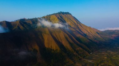 Sembalun 'da, Rinjani Dağı' nın eteklerindeki yuvarlanan tepelerin nefes kesen hava manzarası. Geniş savanalar gün doğumunda altın rengine dönüşüyor..