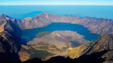 Rinjani 'nin zirvesinden bir hava fotoğrafı aşağıdaki muhteşem Segara Anak Gölü' nü gösteriyor. Canlı mavi gökyüzü ve bulutlarla çevrili gölün dingin güzelliği dramatik manzaraya karşı duruyor..