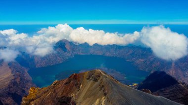 Rinjani 'nin zirvesinden bir hava fotoğrafı aşağıdaki muhteşem Segara Anak Gölü' nü gösteriyor. Canlı mavi gökyüzü ve bulutlarla çevrili gölün dingin güzelliği dramatik manzaraya karşı duruyor..