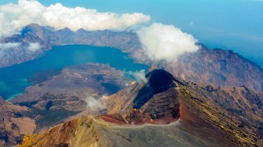 Rinjani 'nin zirvesinden bir hava fotoğrafı aşağıdaki muhteşem Segara Anak Gölü' nü gösteriyor. Canlı mavi gökyüzü ve bulutlarla çevrili gölün dingin güzelliği dramatik manzaraya karşı duruyor..