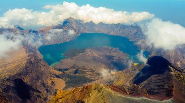 Rinjani 'nin zirvesinden bir hava fotoğrafı aşağıdaki muhteşem Segara Anak Gölü' nü gösteriyor. Canlı mavi gökyüzü ve bulutlarla çevrili gölün dingin güzelliği dramatik manzaraya karşı duruyor..