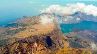 Rinjani 'nin zirvesinden bir hava fotoğrafı aşağıdaki muhteşem Segara Anak Gölü' nü gösteriyor. Canlı mavi gökyüzü ve bulutlarla çevrili gölün dingin güzelliği dramatik manzaraya karşı duruyor..