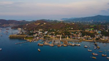 Aerial view of Labuan Bajo harbor at sunset, showcasing numerous pinisi boats docked, with charming local residences surrounding the vibrant scene. clipart