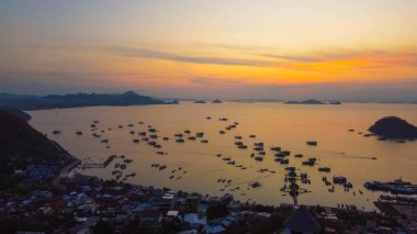 Gün batımında Labuan Bajo limanının hava görüntüsü, çok sayıda pinisi teknesinin kenetlendiğini gösteriyor, büyüleyici yerel konutlar canlı sahneyi çevreledi..