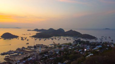 Gün batımında Labuan Bajo limanının hava görüntüsü, çok sayıda pinisi teknesinin kenetlendiğini gösteriyor, büyüleyici yerel konutlar canlı sahneyi çevreledi..