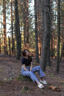 Ormandaki bir kütükte oturan genç bir kadın ağaçların arasından yukarıya bakıyor. Düşünce kavramı, doğa ile bağlantı, huzur ve açık hava yaşam tarzı..