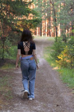 Yeşillik ve yemyeşil ağaçlarla çevrili bir orman yolunda yürüyen genç bir kadının arka görüntüsü. Keşif kavramı, yalnızlık, özgürlük ve doğa ile bağlantı.