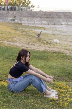 Sarı çiçekli yeşil bir çayırda oturan düşünceli genç kadın arka planda yürüyen kazlar.