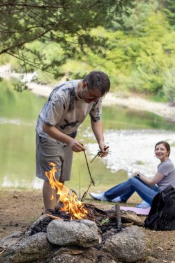 Bir adam ormanda kamp ateşi yakarken bir kadın arka planda oturmuş gülümsüyor. Kamp, aşk, açık hava macerası, hayatta kalma ve birlikte doğanın tadını çıkarma kavramı..