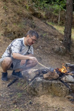 Bir adam ormanda kamp ateşi yakıyor, vahşi doğada sıcak ve samimi bir atmosfer yaratıyor. Kamp, hayatta kalma, açık hava macerası ve doğa bağlantısı kavramı..