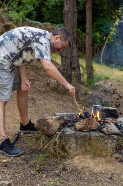 Bir adam ormanda kamp ateşi yakıyor, vahşi doğada sıcak ve samimi bir atmosfer yaratıyor. Kamp, hayatta kalma, açık hava macerası ve doğa bağlantısı kavramı..