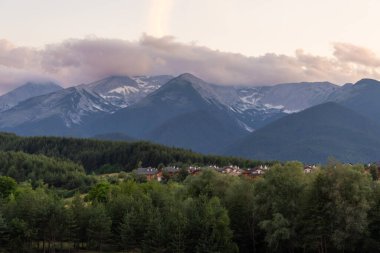Bulgaristan 'ın Bansko kentinde, yemyeşil yemyeşil ile çevrili ahşap, arka planda bulutlu bir gökyüzünün altında yükselen dramatik Pirin dağ zirveleri de var..