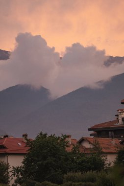 Geleneksel evlerin kısmen kayan bulutlarla kaplı dramatik dağ zirveleriyle buluştuğu Bulgaristan 'ın Bansko kasabasından Pirin Dağları manzarası. Kentsel ve doğal manzaraların uyumlu bir karışımı.