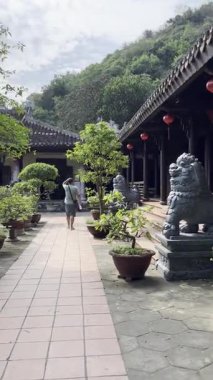 Bir turist, güneşli bir yaz gününde karmaşık mimari, kırmızı fenerler ve sakin dağlarla dolu tarihi bir Vietnam tapınağı kompleksini fotoğraflıyor. (158 karakter). Mermer Dağlar 