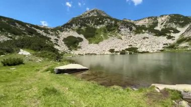 Sakin bir dağ gölü, Bulgaristan 'ın Pirin kentinin berrak mavi gökyüzünü ve engebeli tepelerini yansıtıyor. Parlak yaz güneşinin altında yemyeşil çimenler ve çam çalıları sakin suları kaplıyor..