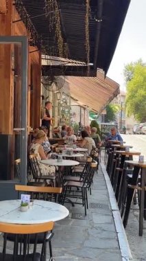 Güneşli bir yaz gününde insanlar açık bir kafede kahve içip sohbet etmekten zevk alıyorlar. Kafede bir tente, sicim lambaları ve güzel bir kaldırıma dökülen masalar var..