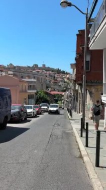 Yunanistan 'ın Kavala kentinde canlı bir sokak sahnesi, tarihi Bizans kalesi ve antik Kamares su kemerine doğru yokuş yukarı gidiyor. Arabalar ve yayalar açık mavi bir yaz gökyüzünün altında hareket ederler..