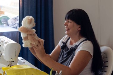 Gülümseyen bir kadın, parlak bir atölyede neşeli zanaatkarlığını sergileyen, el yapımı, koleksiyonluk bir oyuncak ayıyı gururla elinde tutuyor. Uzun siyah saçları omuzlarının üzerinden akıyor. Yaratılışına hayranlık duyuyor..