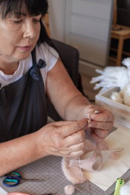 Odaklanmış bir kadın zarif pembe bir tekstil eşyasını, muhtemelen örgü ya da dokuma işine dalmış durumda. Elleri, atölyesinde çeşitli el aletleri ile çevrili ipliği dikkatlice manipüle ediyor..