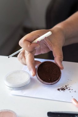 Eller kozmetik ürünlerini hazırlar: bir fırça tutarken, koyu kahverengi barut kabı açıkken. Beyaz toz kabı beyaz bir yüzeye döküldü.