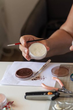 Bir sanatçının elleri bir tencere toz ve küçük bir fırça tutmakla meşguldür. Çeşitli pigmentler, fırçalar ve el sanatları araçları serilmiş, titiz bir yaratıcı projeye işaret ediyor..