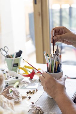 Bir tutucudan boya fırçası seçerken yakın plan, çeşitli el yapımı malzemelerle çevrili, boncuklar, kumaşlar, ve parlak bir masanın üzerindeki aletler, yaratıcı bir proje için hazır..
