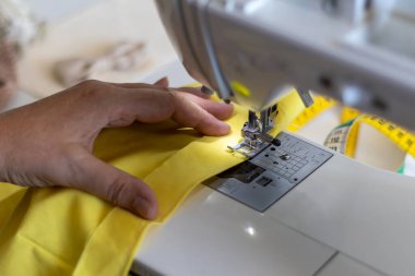 Dikiş makinesinde çalışan bir terzinin parlak sarı malzemelerle yeni bir parça yarattığının detaylı görüntüsü. El yapımı projelerdeki hassasiyet ve adanmışlığı vurguluyor.