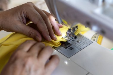 Yakın plan: Eller, dikiş makinesi iğnesinin altında parlak sarı kumaşa ustalıkla rehberlik eder, terzilik hassasiyetini gösterir.