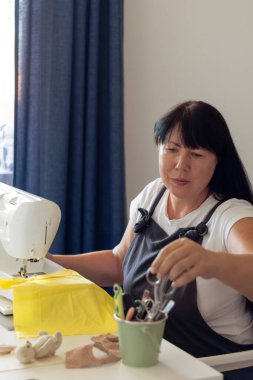 Önlük giymiş odaklanmış bir kadın dikiş makinesi üzerinde çalışıyor, el yapımı eşyalar yapıyor. El sanatları hobisini sergilemek için bir kova aletten makas alıyor..