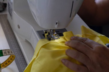 Eller, canlı sarı kumaşlı yeni bir parça yaratmak için bir dikiş makinesi üzerinde özenle çalışır. Yakınlarda zanaatın hassasiyetini vurgulayan bir mezura var..