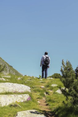 Sırt çantalı bir adam Pirin Ulusal Parkı 'ndaki açık mavi gökyüzünün altında manzaralı, çimenli bir dağ yolunda yürüyor. Görüntü macerayı, keşfi ve doğanın dingin güzelliğini çağrıştırıyor..