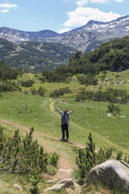 Dolambaçlı bir yolda neşeli bir yürüyüşçü, kollarını açmış Pirin Ulusal Parkı 'nın nefes kesen doğal güzelliğini kutluyor. Görkemli karlı dağlar ve yemyeşil vadiler manzarayı çerçeveliyor.