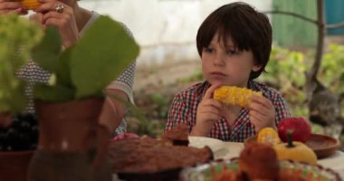 Okul çağındaki çocuk elinde mısır koçanı tutuyor, ısırıyor, çiğniyor ve ızgara mısır yiyor. Çocuk mısır koçanı yiyor, annesinin yanında oturuyor ve sohbet ediyor. Aile yemeği.