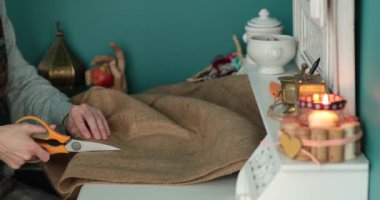 Kadın beyaz masada büyük bir makasla çuval bezi kesti. Mum ve klasik süslemelerle. El işi çuval bezi kumaşı, ev yapımı ürünleri dikmek için biyolojik olarak ayrıştırılabilir el işi malzemesi kullanılıyor. El işi köşesi