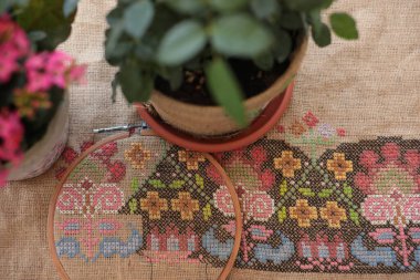 Traditional cross stitched folk motif embroidery on natural burlap fabric background closeup on foreground with houseplants in flower pots on blurred foreground. Needlework embroidery crafting hobby clipart