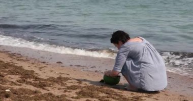 Sahilde tanınmayan bir kadın deniz suyuyla midyeleri kasede yıkıyor ve kötü olanları suya atıyor. Kadın deniz suyunda kumun üzerinde oturuyor. Dalgalar kıyıya vurur, dalgaların köpüğü