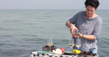 Kadın şarap kadehlerine beyaz şarap döker, sonra iki orta yaşlı kadın tezahürat yapar ve deniz suyu arka planında şarap içerler. Deniz ürünleri midyeli masa. Deniz kenarında yaz pikniği