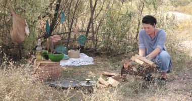Kadın yemek pişirmek için ormanda kamp ateşi yakıyor. Kütükler birbiri üzerine konur, ateş çıkar. Şenlik ateşinde yemek pişirmek. Yemek için yemek takımları, malzemeler, ürünler hazırlayın. Yazın açık hava bbq pikniği