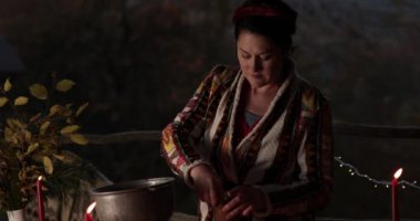 Geleneksel desenli elbiseli bir kadın, yılbaşı pazarında, bulanık Noel ışıkları ve yanan mumlarla şarabı sulandırdı ve tarçın kattı. 40 'lı yaşlardaki kadınlar sıcak bayram içkisi yapar.