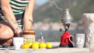 Dövmeli bir kadın, Karadağ 'ın Donji Stoliv körfezindeki bir rıhtımda piknik yaparken gaz ocağında kaşık alıp sıcak kahve karıştırıyor. Kadın, arka planda bulanık dağ manzaralı açık havada kahve yapıyor.